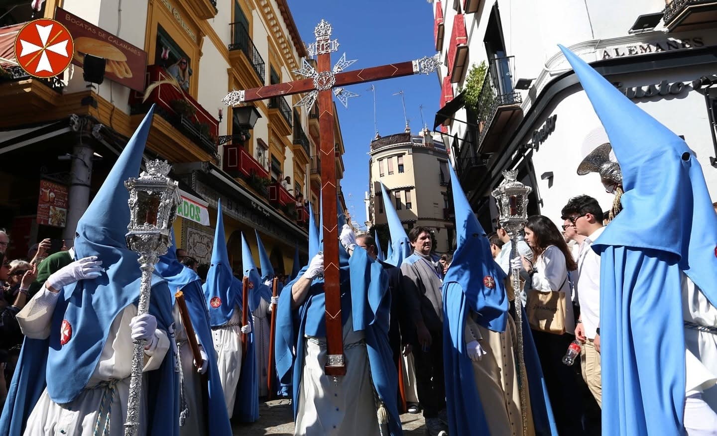 Cruz de guía y nazarenos de túnica crema y antifaz celeste de la Hermandad de San Esteban avanzando entre el público.